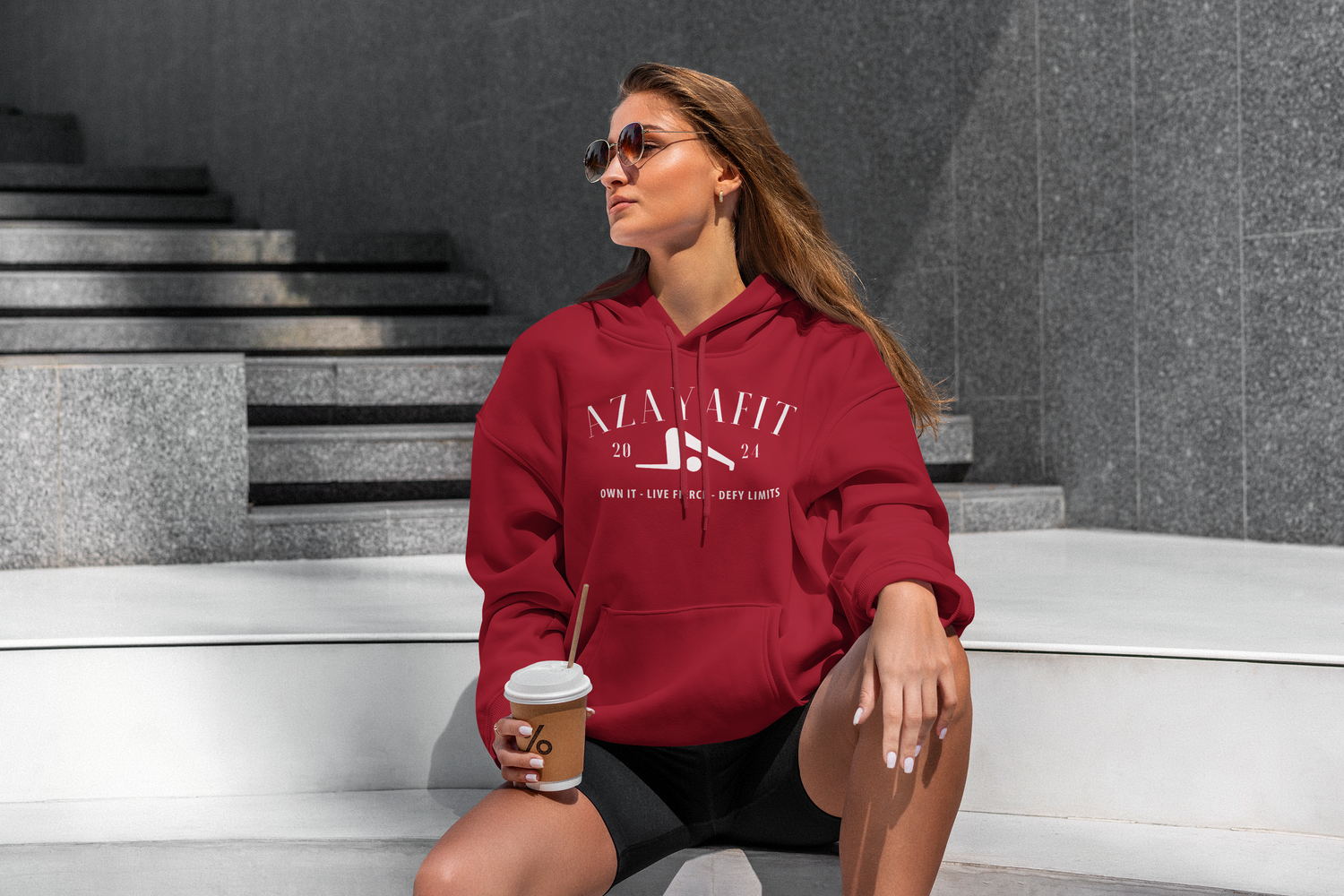 Woman wearing a red hoodie with a brand logo, sitting on steps holding a coffee cup.