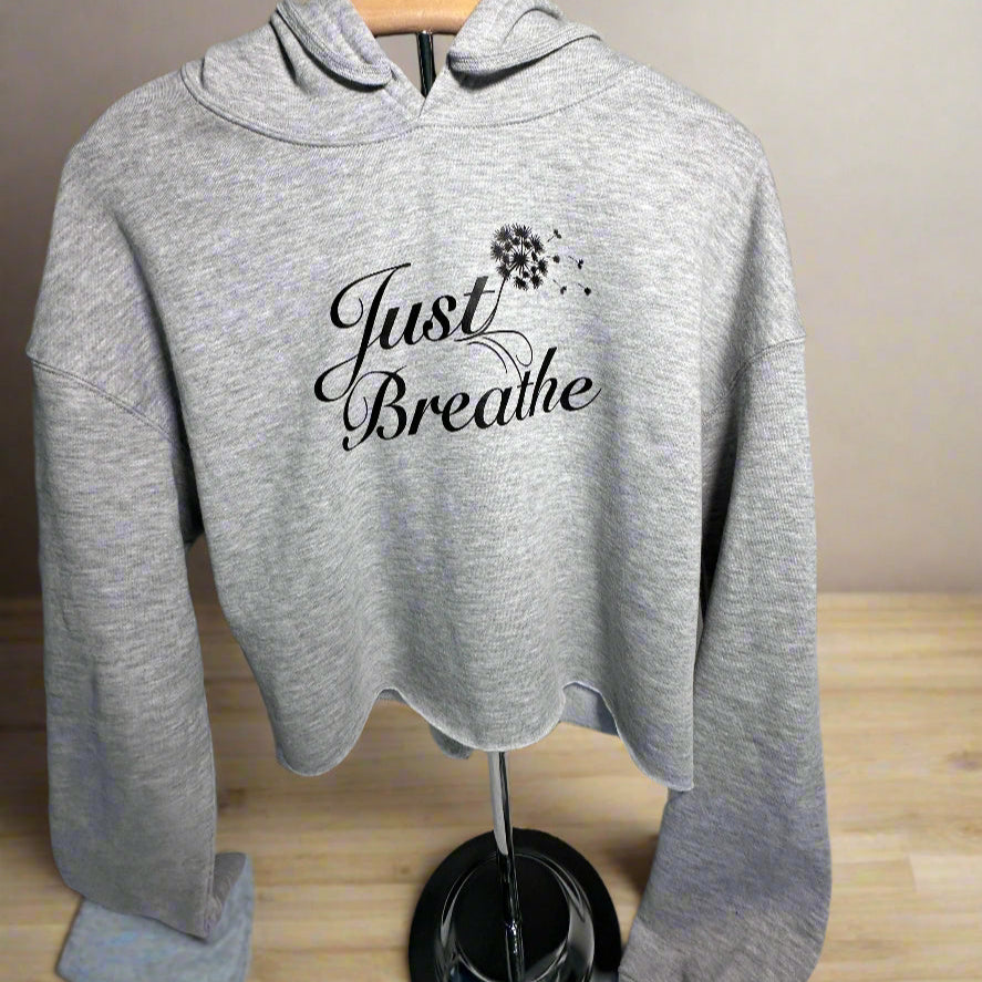 Heather grey cropped hoodie displayed on a stand atop a table, featuring the elegant "Just Breathe" design with a delicate dandelion blowing from the "T" in "Just." The setup highlights the hoodie’s soft fabric and stylish aesthetic.