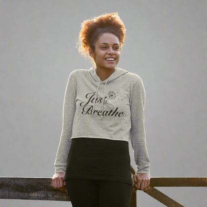 Happy woman leaning on a wooden fence while wearing the heather grey Just Breathe crop hoodie, a stylish and comfortable choice for casual wear and workouts.