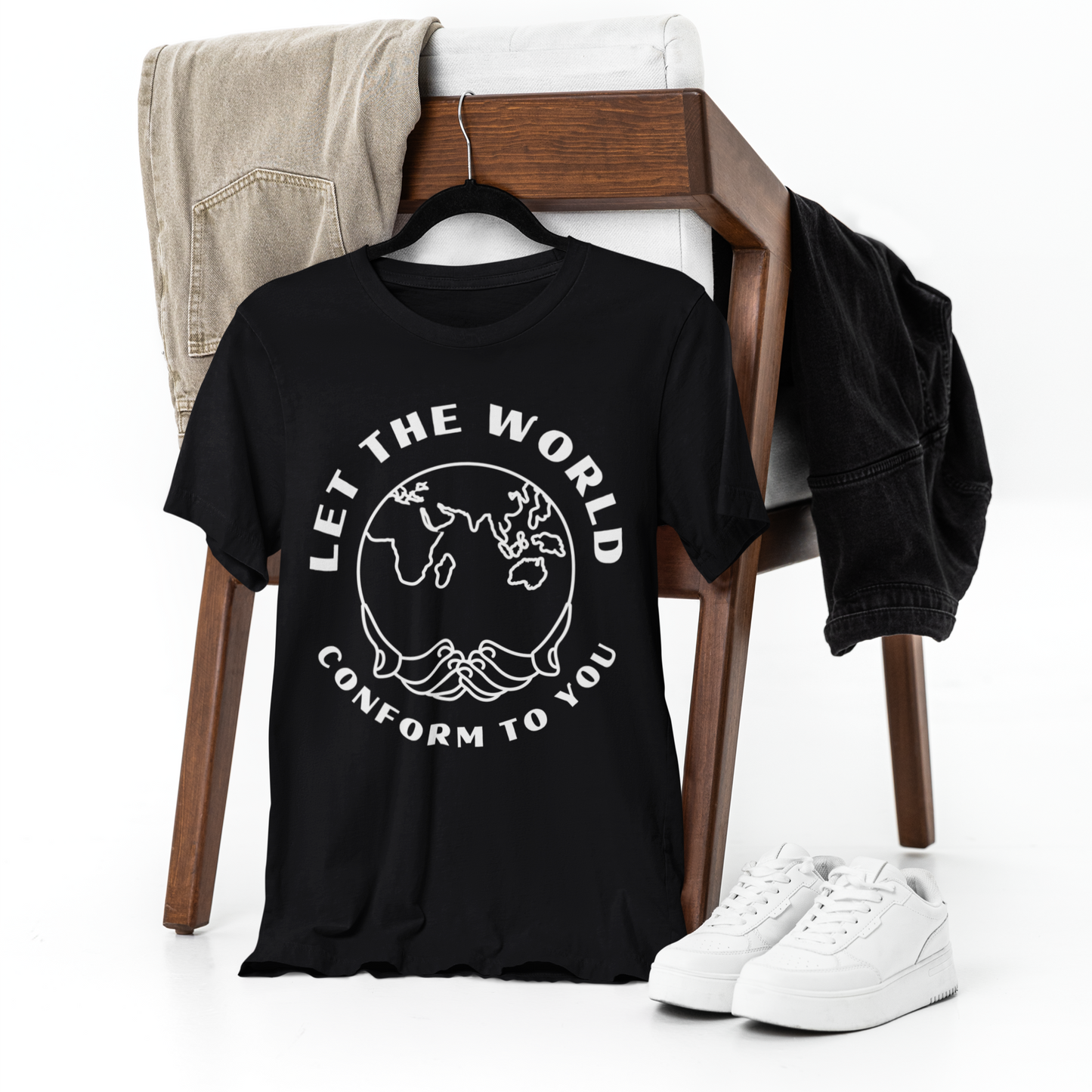 Black T-shirt featuring the "Let the World Conform to You" design draped over a chair, with matching pants casually placed over the backrest. A pair of white shoes rests on the ground beside the chair, all set against a clean white backdrop.