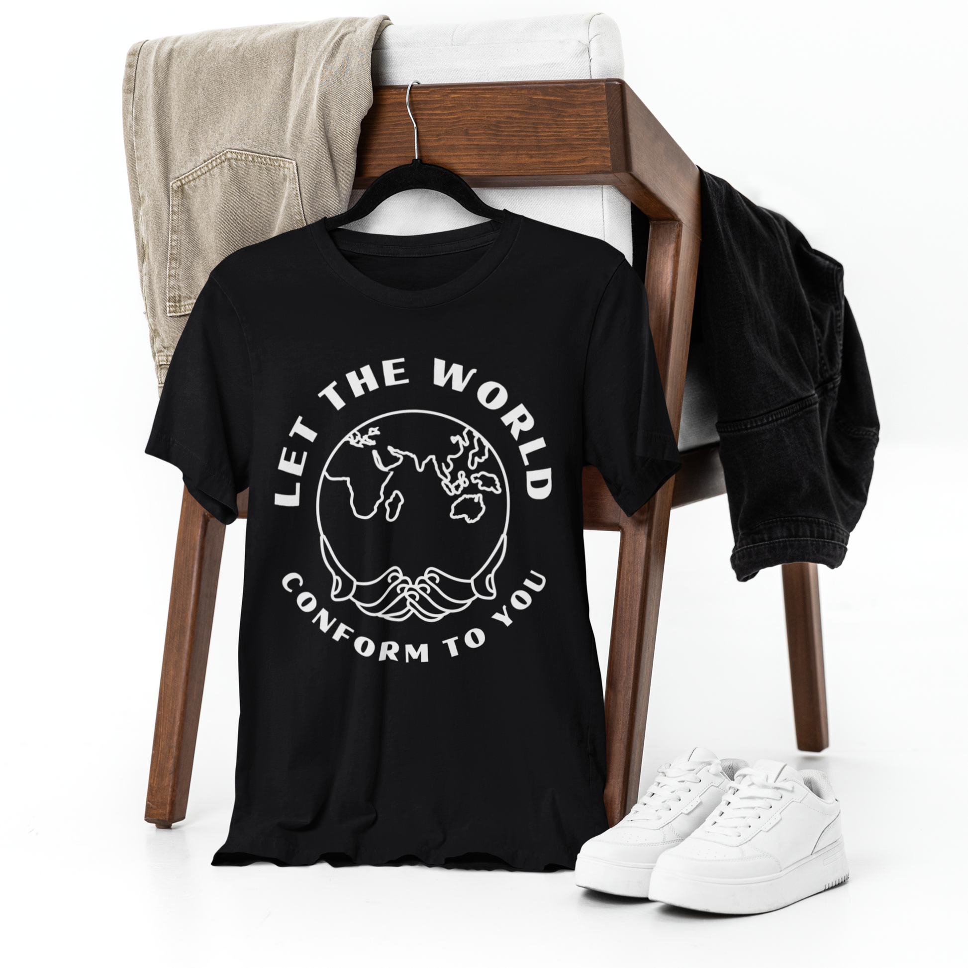 Black T-shirt featuring the "Let the World Conform to You" design draped over a chair, with matching pants casually placed over the backrest. A pair of white shoes rests on the ground beside the chair, all set against a clean white backdrop.