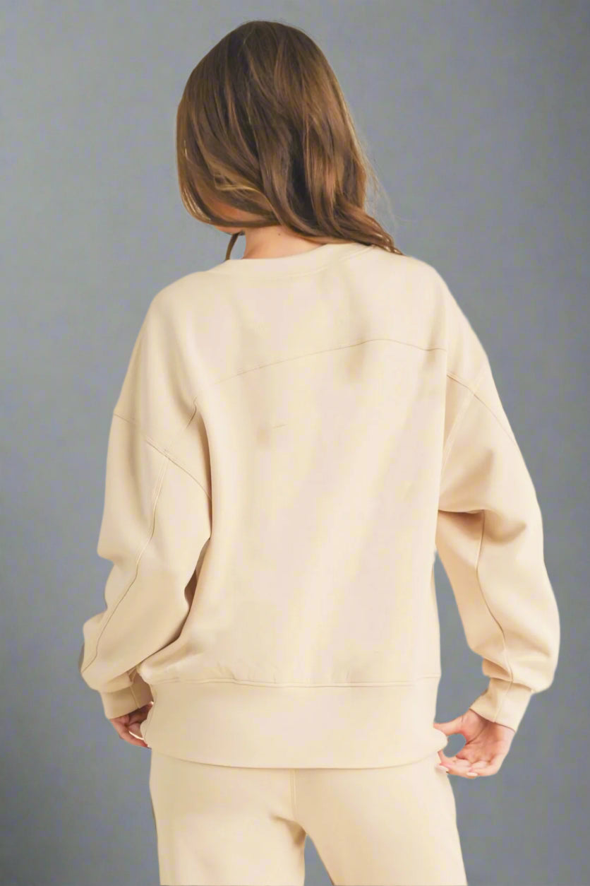 Model standing with back facing the camera against a grey backdrop, wearing an oat-colored sweatshirt and matching sweatpants. The relaxed fit and soft fabric are emphasized, highlighting the casual and cozy design.