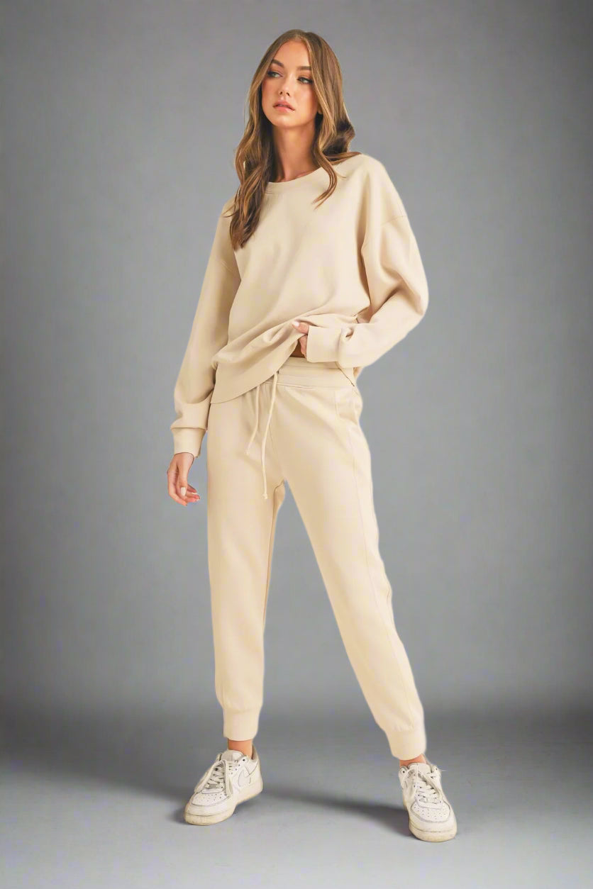 Model posing in a full-length front view against a grey backdrop, wearing an oat-colored sweatshirt and matching sweatpants. The outfit showcases a relaxed fit and neutral tone, perfect for casual comfort.
