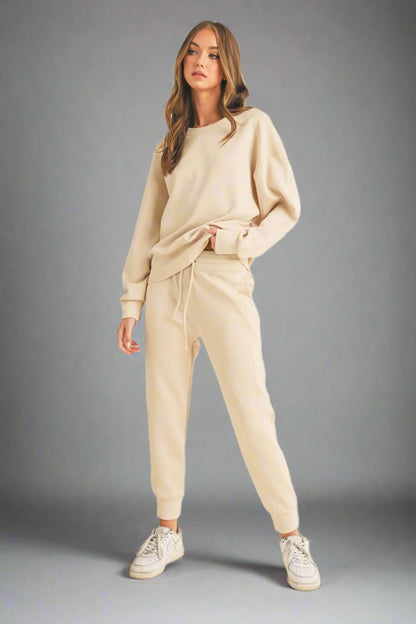 Model posing in a full-length front view against a grey backdrop, wearing an oat-colored sweatshirt and matching sweatpants. The outfit showcases a relaxed fit and neutral tone, perfect for casual comfort.