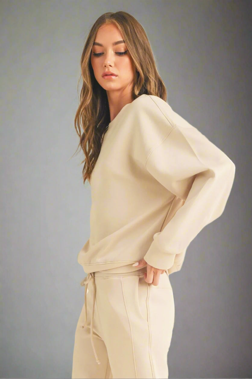 Model posing in a side view against a grey backdrop, wearing an oat-colored sweatshirt and matching sweatpants. The relaxed fit and cozy fabric are highlighted, showcasing the comfortable loungewear set.