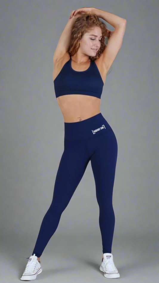 Full-length image of a woman posing with both hands crossed above her head, wearing royal blue full-length leggings with "Radiant Vibe" printed in small white letters on the left hip. She pairs it with a matching sports bra, showcasing a stylish and coordinated activewear set.