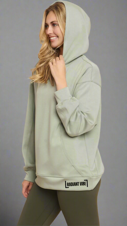 Cropped image of a model wearing a sage green hoodie with "Radiant Vibe" printed in small black letters on the left hip area. The model is posing from the side with the hood up, highlighting the fit and design details of the hoodie.