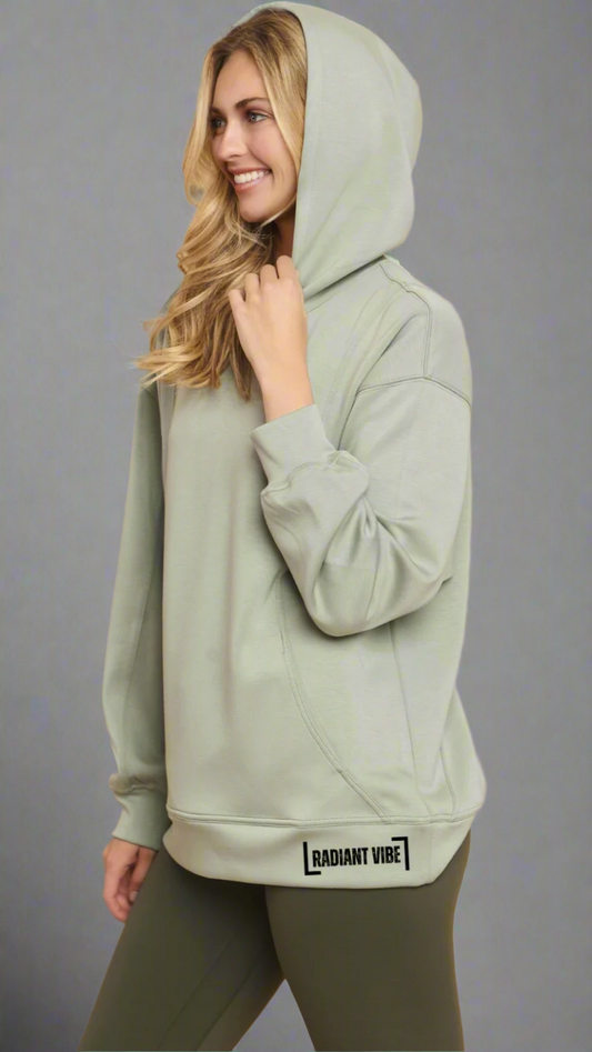Cropped image of a model wearing a sage green hoodie with "Radiant Vibe" printed in small black letters on the left hip area. The model is posing from the side with the hood up, highlighting the fit and design details of the hoodie.