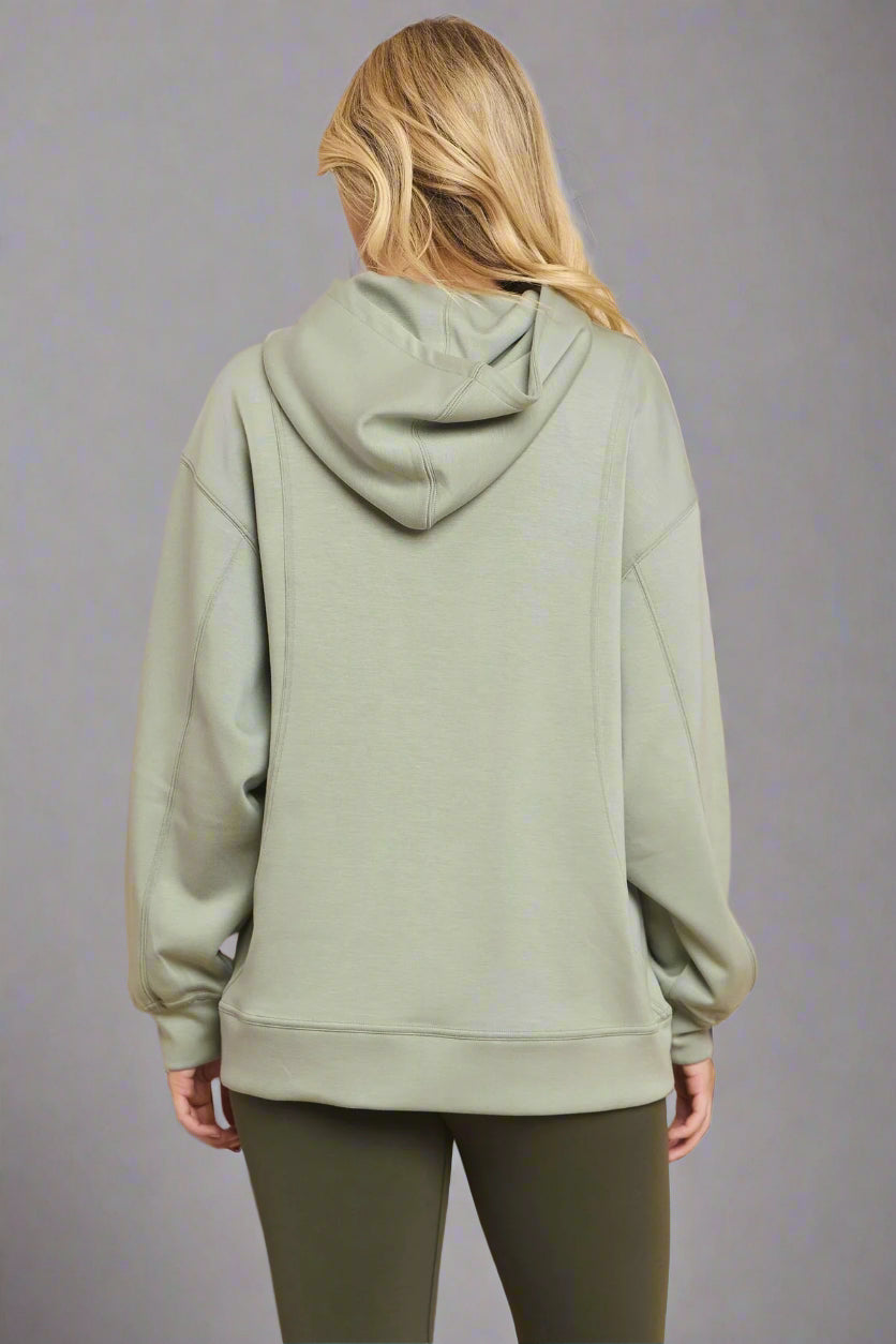 Backside view of a model wearing a sage green hoodie against a grey backdrop. The relaxed fit and soft fabric are highlighted, showcasing the hood and overall design.