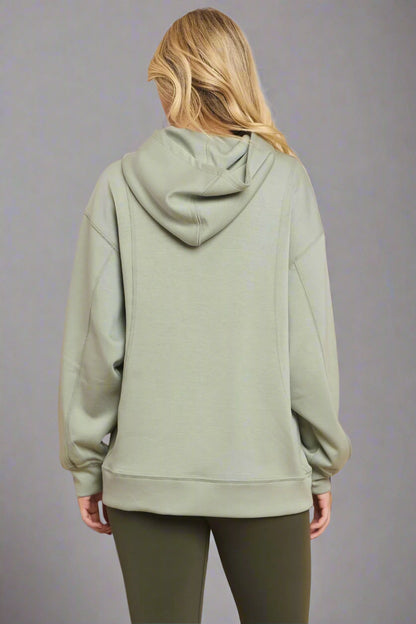 Backside view of a model wearing a sage green hoodie against a grey backdrop. The relaxed fit and soft fabric are highlighted, showcasing the hood and overall design.