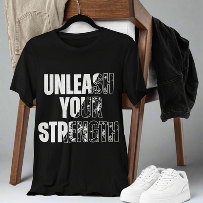 Black T-shirt with "Unleash Your Strength" design draped over a chair, paired with pants and white shoes on the ground. The setup is against a white backdrop, emphasizing the bold black and white lettering of the design.